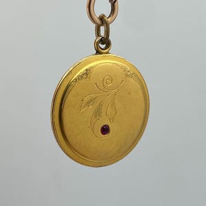 Könnte beinhalten: Ein goldfarbenes, rundes Medaillon mit floralem Design und einem kleinen, roten Edelstein. Das Medaillon ist an einer goldenen Kette und einem Verschluss befestigt. Die Oberfläche des Medaillons hat ein eingraviertes Blumenmuster und einen kleinen, runden, roten Edelstein.