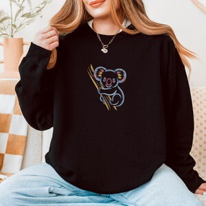 May include: Black sweatshirt featuring a colorful embroidered koala bear clinging to a branch. The koala has pink, blue, and yellow accents. The sweatshirt is a solid black color and has a crew neck.