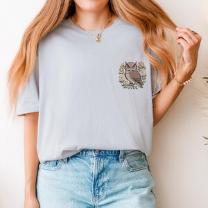 May include: Light blue t-shirt featuring an embroidered owl design. The owl is brown and beige, surrounded by a floral wreath. The shirt is a soft, casual style, suitable for everyday wear.