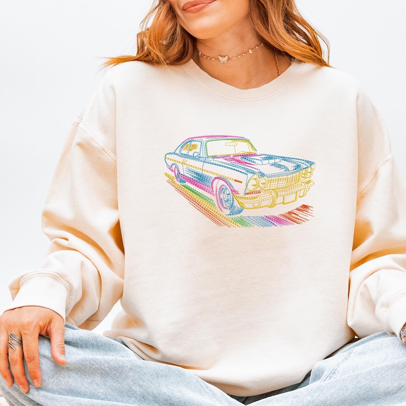 May include: Cream-colored sweatshirt featuring a colorful, stylized car graphic. The car design incorporates a rainbow of colors, including pink, blue, yellow, and green. The sweatshirt has long sleeves and a crew neck.