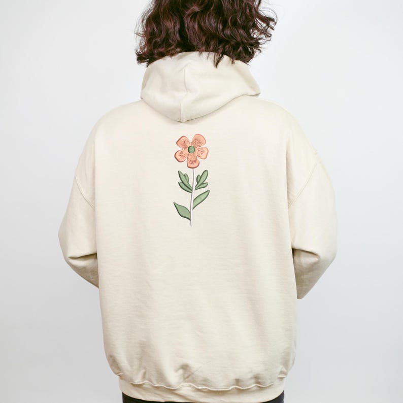 May include: A cream-colored hoodie with a floral design on the back. The flower is peach-colored with a green stem and leaves. The hoodie has a hood and long sleeves.