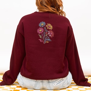 May include: A burgundy sweatshirt with an embroidered floral design on the back. The floral design features blue, yellow, pink, and purple flowers with green leaves. The sweatshirt has long sleeves and a crew neck.