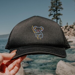 May include: Black trucker hat with a mesh back and a curved brim. The hat features an embroidered koala bear holding a branch, with colorful accents. The hat is held by a hand, with a scenic background of water and trees.