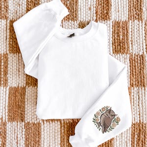 May include: White sweatshirt with an owl design on the left sleeve. The owl is brown and tan, surrounded by a floral wreath in shades of brown and green. The sweatshirt is made of a soft material and is perfect for casual wear.