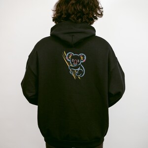 May include: Black hoodie featuring a colorful embroidered koala holding a branch. The koala has blue, pink, and yellow accents. The hoodie has a drawstring hood and a kangaroo pocket.