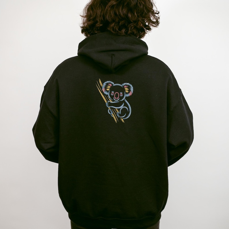 May include: Black hoodie featuring a colorful embroidered koala holding a branch. The koala has blue, pink, and yellow accents. The hoodie has a drawstring hood and a kangaroo pocket.