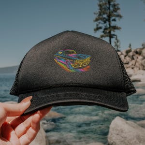 May include: Black trucker hat with a mesh back and a curved brim. The front of the hat features a colorful embroidered design of a classic car. The car is outlined in bright colors, including blue, yellow, and pink.