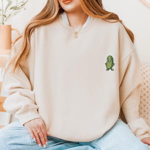 Może przedstawiać: Beżowa bluza z małym, haftowanym, kresk&oacute;wkowym og&oacute;rkiem z przodu. Og&oacute;rek ma uśmiechniętą twarz i zielone buty. Bluza jest w neutralnym kolorze i ma okrągły dekolt.