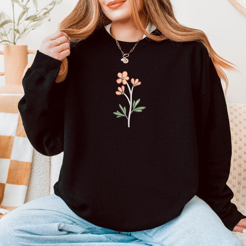 May include: Black sweatshirt featuring a floral design with peach-colored flowers and green leaves. The sweatshirt has a crew neck and long sleeves. The floral design is centered on the chest.