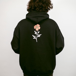 May include: Black hoodie with a floral embroidery on the back. The embroidered flower has pink petals, a green center, and white leaves and stem. The hoodie has a hood and a relaxed fit.