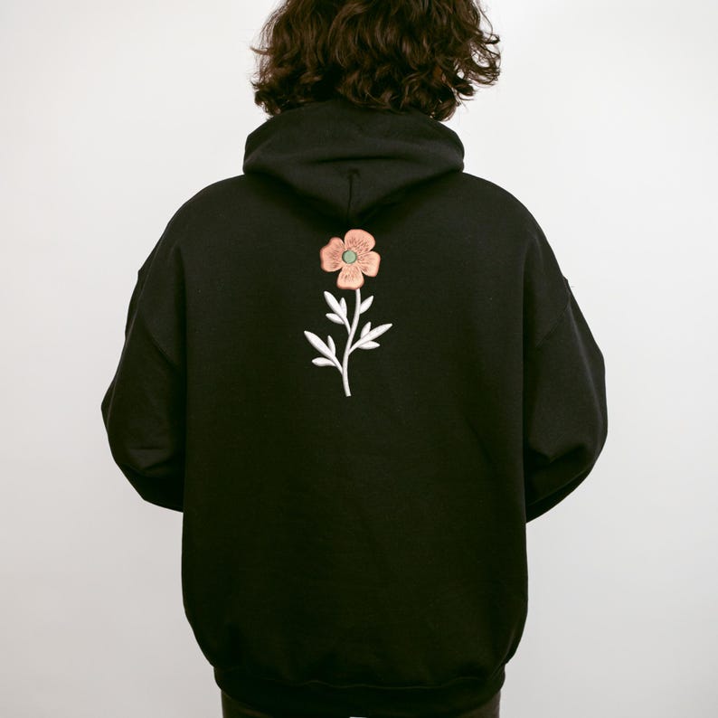 May include: Black hoodie with a floral embroidery on the back. The embroidered flower has pink petals, a green center, and white leaves and stem. The hoodie has a hood and a relaxed fit.