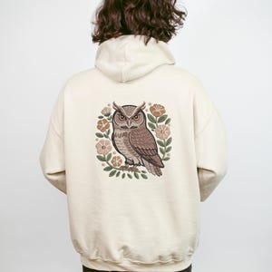May include: Cream-colored hoodie featuring a detailed illustration of an owl surrounded by flowers. The owl is brown and tan, with large eyes and a focused gaze. The floral elements are in shades of beige and green, creating a nature-inspired design.