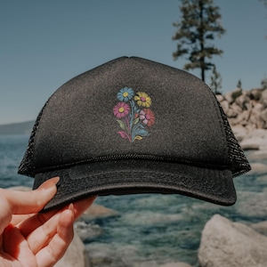 May include: Black trucker hat featuring a colourful floral embroidery. The embroidered flowers have blue, yellow, pink, and red petals with green stems and leaves. The hat has a mesh back and a curved peak.