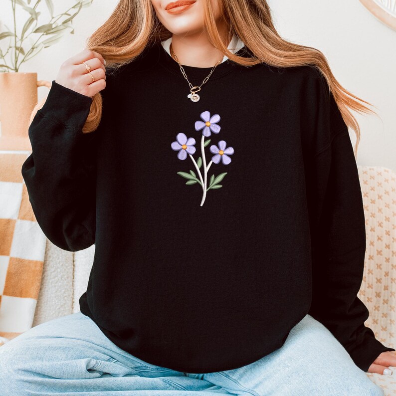 May include: Black sweatshirt featuring a floral design. The graphic shows three lavender-colored flowers with green leaves and a white stem. The sweatshirt has a crew neck and long sleeves, suitable for casual wear.