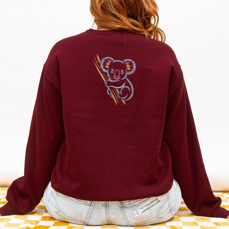 May include: Burgundy sweatshirt featuring a colorful embroidered koala clinging to a branch. The koala has blue, pink, and yellow accents. The sweatshirt is a long-sleeved crewneck style. The person wearing the sweatshirt is sitting on a yellow and white checkered surface.