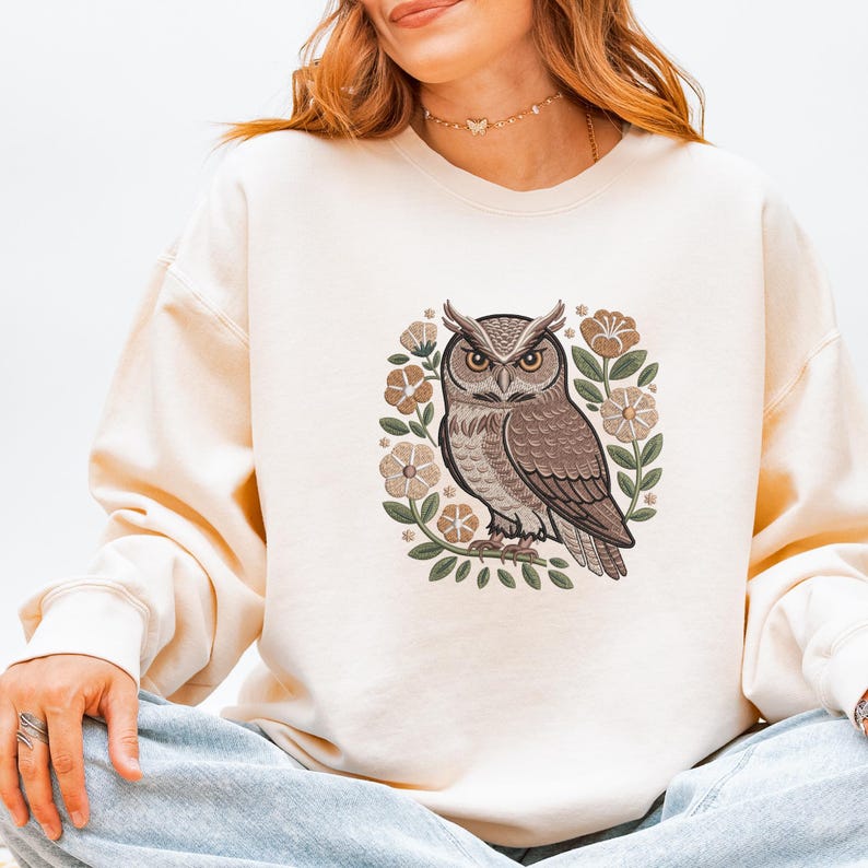 May include: Cream-colored sweatshirt featuring an embroidered owl design. The owl is brown and tan, surrounded by tan and green floral elements. The sweatshirt has long sleeves and a crew neck.