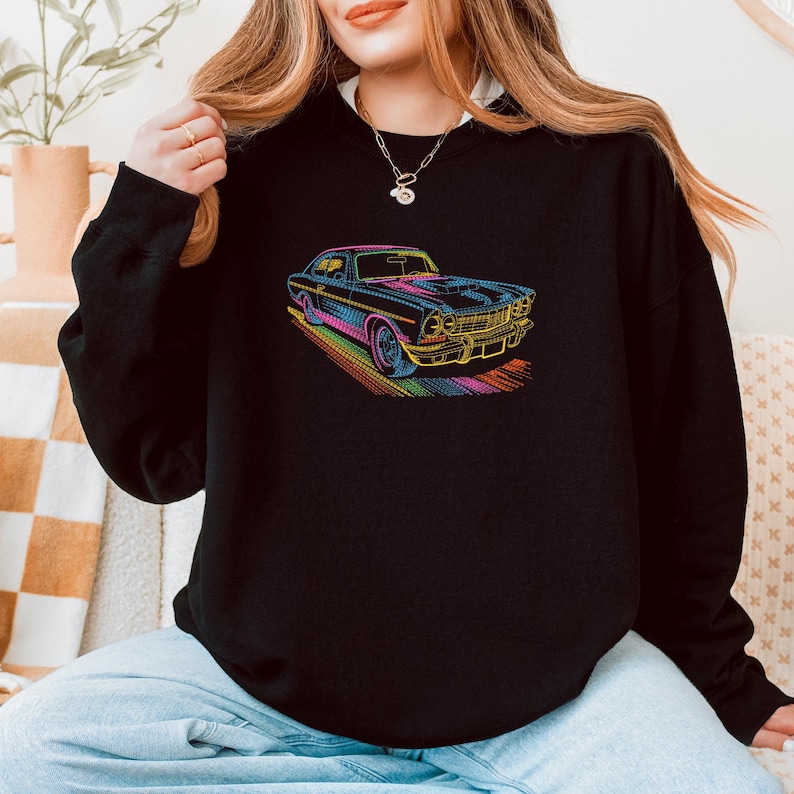 May include: Black sweatshirt featuring a colorful, neon-outlined illustration of a classic car. The car design includes details like the grille, headlights, and wheels, with a rainbow-colored trail beneath it. The sweatshirt is a solid black color.