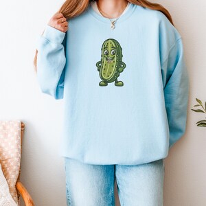 Może przedstawiać: Jasnoniebieska bluza z nadrukiem postaci z kresk&oacute;wki - og&oacute;rka. Zielony og&oacute;rek z ż&oacute;łtymi detalami uśmiecha się i ma ręce i nogi. Bluza wykonana jest z miękkiego materiału i ma wygodny kr&oacute;j.