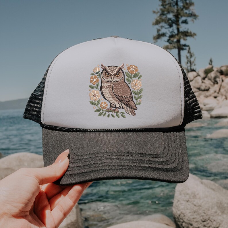 May include: A trucker hat with a white front panel featuring a brown owl illustration surrounded by flowers and leaves. The hat has a gray brim and a black mesh back. The owl design is detailed with brown and tan colors.