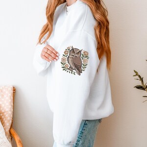 May include: White sweatshirt featuring an embroidered owl design. The owl is brown and tan, perched on a branch, surrounded by a wreath of flowers and leaves. The sweatshirt is a casual, comfortable style.