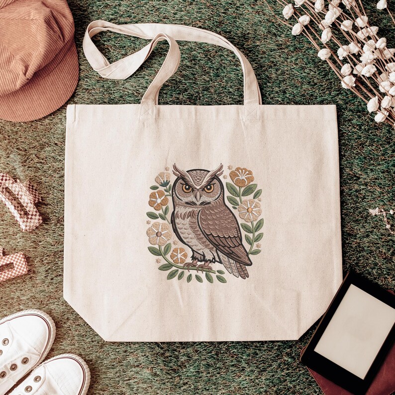May include: Cream-colored tote bag featuring a detailed illustration of a brown owl surrounded by flowers and leaves. The owl has yellow eyes and is centered on the bag. The bag has two cream-colored handles.