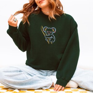 May include: Dark green sweatshirt featuring a colorful embroidered koala clinging to a branch. The koala has a light blue body, pink ears, and yellow accents. The sweatshirt is a solid color with long sleeves.