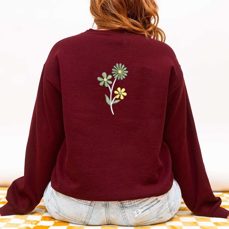 May include: A maroon sweatshirt with a floral embroidery design on the back. The embroidered flowers are in shades of yellow and green, with a white stem. The sweatshirt has long sleeves and a crew neck.