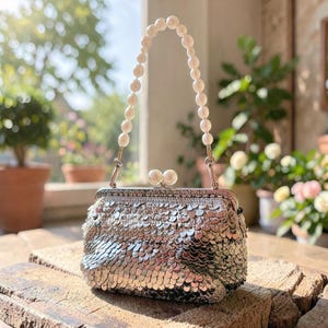 May include: A silver sequined handbag with a pearl beaded handle. The bag has a clasp closure and is embellished with shiny sequins. The handle is made of white pearls. The bag is on a wooden surface.