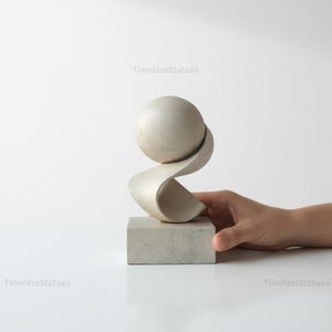 May include: An abstract sculpture in light grey, featuring a sphere balanced on a flowing, ribbon-like shape, set on a square base. The text "TimelessStatues" is visible. A modern art piece.