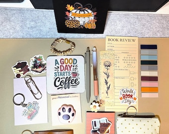 May include: A collection of stationery items and accessories, such as pens, stickers, a scrunchie, a keychain, and a small coin purse. A black box with a pumpkin design is at the top. A book review card and sticky notes are also visible.