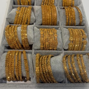 May include: A collection of gold-coloured metal bangles arranged in sets on grey velvet cushions within a display tray. Each set features distinct intricate textures, patterns, and link designs, showcasing a variety of metallic finishes and shapes.