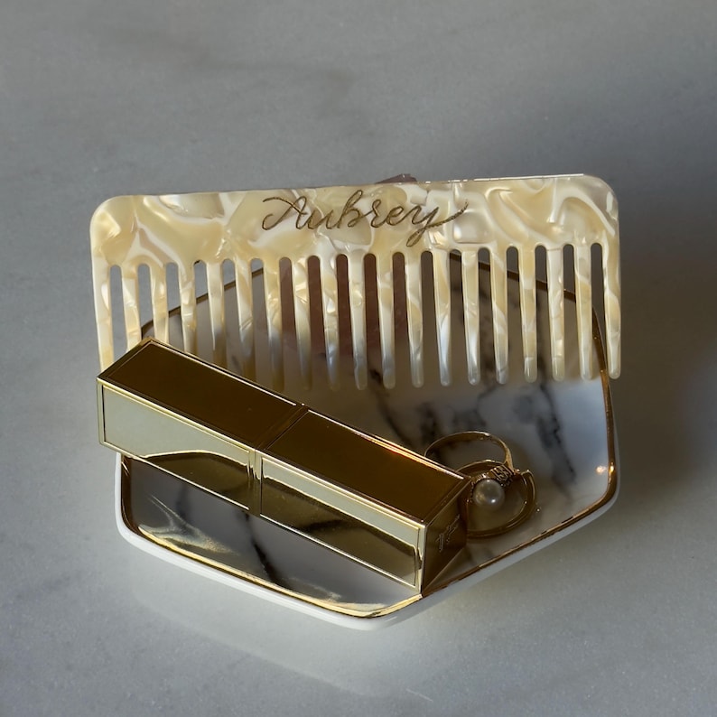 May include: A vanity set with a cream-coloured comb, gold lipstick, and a ring in a small dish. The comb is inscribed with "Aubrey" in gold script. The dish has a marble pattern and gold trim. The lipstick is gold-toned.