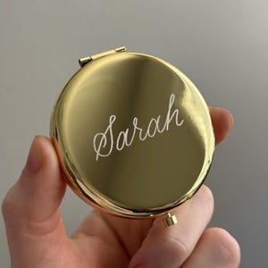 May include: A round, gold-colored metal compact mirror with a polished finish. The word Sarah is engraved in a white cursive script across the center of the lid. The compact features a small hinge at the top and a small latch at the bottom.