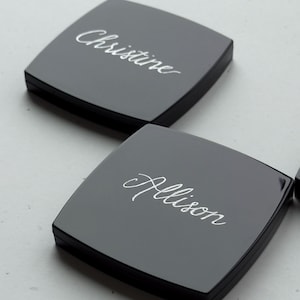 May include: Two square, black compacts with rounded corners. The name "Christine" is elegantly inscribed in white script on one compact, and "Allison" on the other. The compacts are set against a light background.