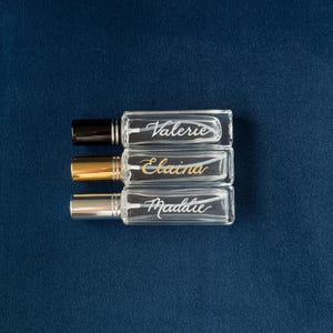 May include: Three rectangular glass perfume bottles with silver, gold, and black caps are arranged vertically. Each bottle is labeled with a name: Valerie, Elaina, and Maddie, in elegant script. The bottles rest on a dark blue surface.