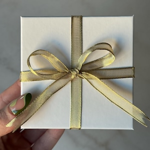 May include: A square, white gift box tied with a gold ribbon in a cross pattern and a bow. The ribbon has a subtle sheen. The box is held in a hand with a painted nail.