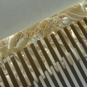 May include: Ivory-coloured comb with the name "Aubrey" in gold script. The comb has a pearlescent finish and a series of evenly spaced teeth. The comb is angled, casting shadows across the teeth.