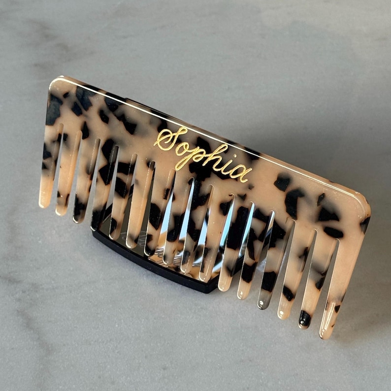 May include: A tortoiseshell-patterned hair comb with the name "Sophia" in gold script. The comb has a rectangular shape with evenly spaced teeth and a black base. The comb is designed for styling and grooming hair.