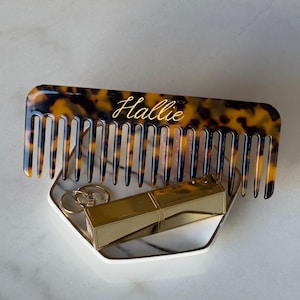 May include: A tortoiseshell comb with the name "Hallie" in gold script rests on a marble tray with gold lipstick and rings. The comb's teeth are evenly spaced. The tray is white with a gold trim.