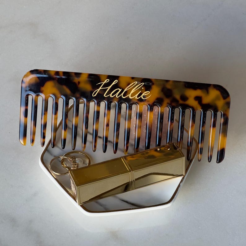 May include: A tortoiseshell comb with the name "Hallie" in gold script rests on a marble tray with gold lipstick and rings. The comb's teeth are evenly spaced. The tray is white with a gold trim.