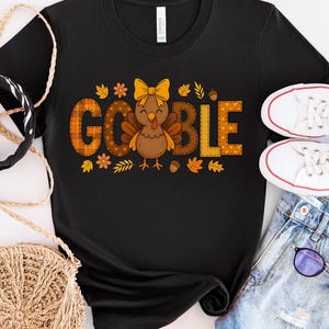 May include: Black t-shirt with a Thanksgiving design. The word "GOBBLE" is in orange and brown plaid letters, with a cartoon turkey in the middle. The design includes autumn leaves, flowers, and acorns. White trainers and denim shorts are also visible.
