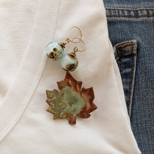 May include: A pair of earrings with blue and brown glass beads and a ceramic leaf charm. The earrings are hanging on a white t-shirt.
