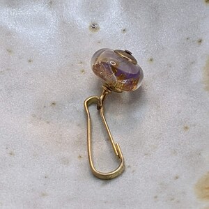 May include: A gold-toned charm with a clear glass bead containing purple and gold flecks. The charm is attached to a gold-toned clasp.