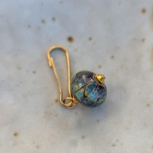 May include: A gold safety pin earring with a blue and green glass bead. The bead has a gold top and a textured surface.
