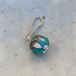 May include: A teal blue glass bead with white and brown swirls, attached to a silver metal lobster clasp.