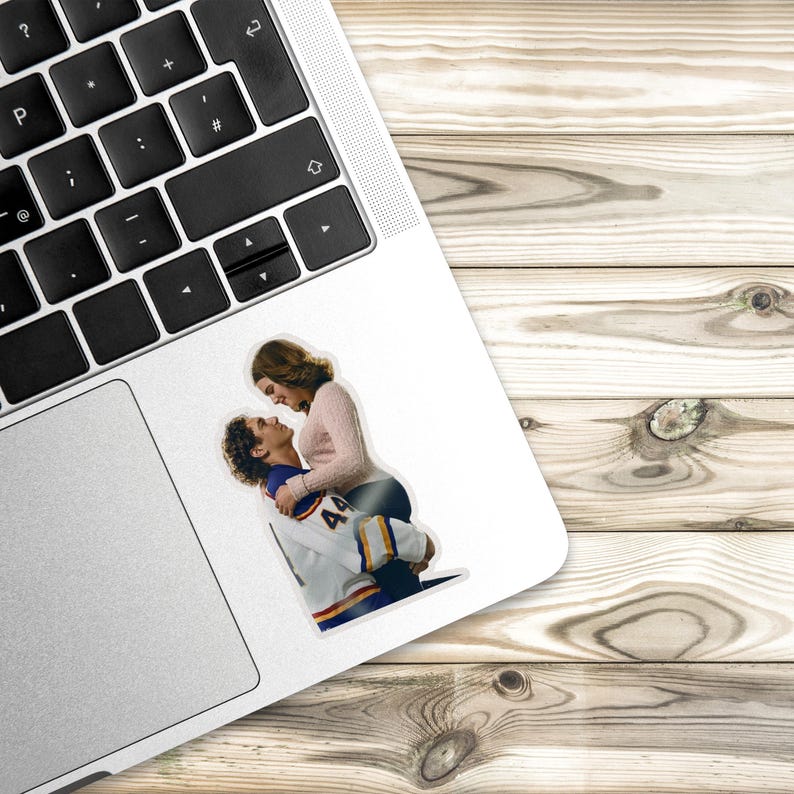 May include: A die-cut sticker featuring a person with brown hair wearing a light pink long-sleeved top embracing another person wearing a white, blue, and yellow hockey jersey with the number 44 on the sleeve, applied to a laptop surface.