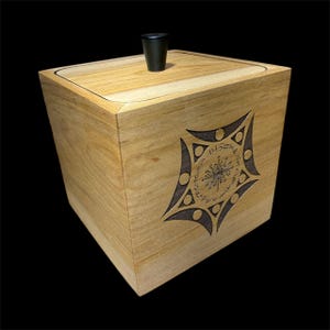 May include: A wooden box with a light brown finish and a black knob on the lid. The box features a dark, stylized geometric design on the front. The design is a star-like shape with circular and triangular elements.