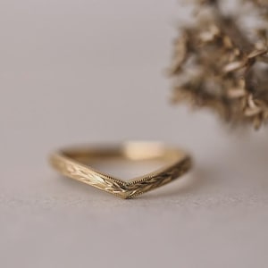 May include: A gold-coloured chevron ring with an engraved leaf pattern. The ring has a V-shape design and is set against a soft, neutral background. The ring is a piece of jewellery.