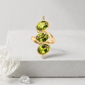 Puede incluir: Un anillo de oro con tres piedras preciosas de peridoto de corte ovalado alineadas verticalmente, cada una acentuada con una pequeña piedra preciosa redonda de color rosa intenso engastada en el lateral del bisel. La banda es lisa y pulida.