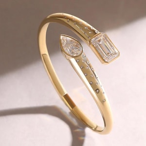 Könnte beinhalten: Ein goldfarbenes Armband mit offenem Design. Es hat einen birnenförmigen Diamanten und einen Diamanten im Smaragdschliff. Kleine, runde Diamanten sind entlang des Armbands gesetzt. Ein elegantes Schmuckstück.